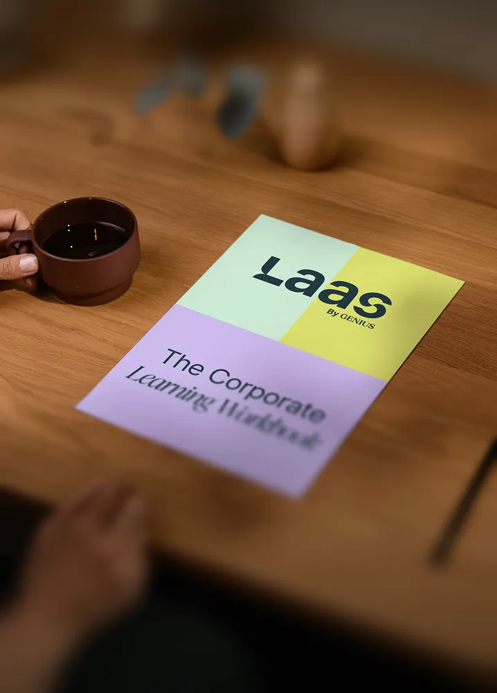 Hand holding a brown coffee cup next to a colorful brochure titled 'Laas by Genius: The Corporate Learning Workbook' on a wooden table.