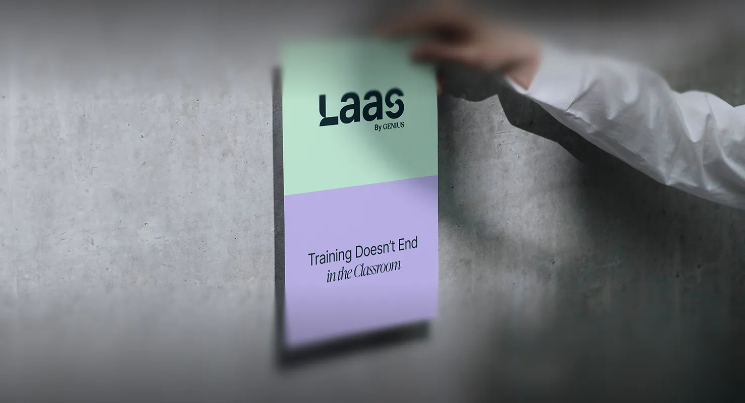 Hand pinning a green and purple sign on a gray wall that reads 'Laas By GENIUS' and 'Training Doesn’t End in the Classroom'.