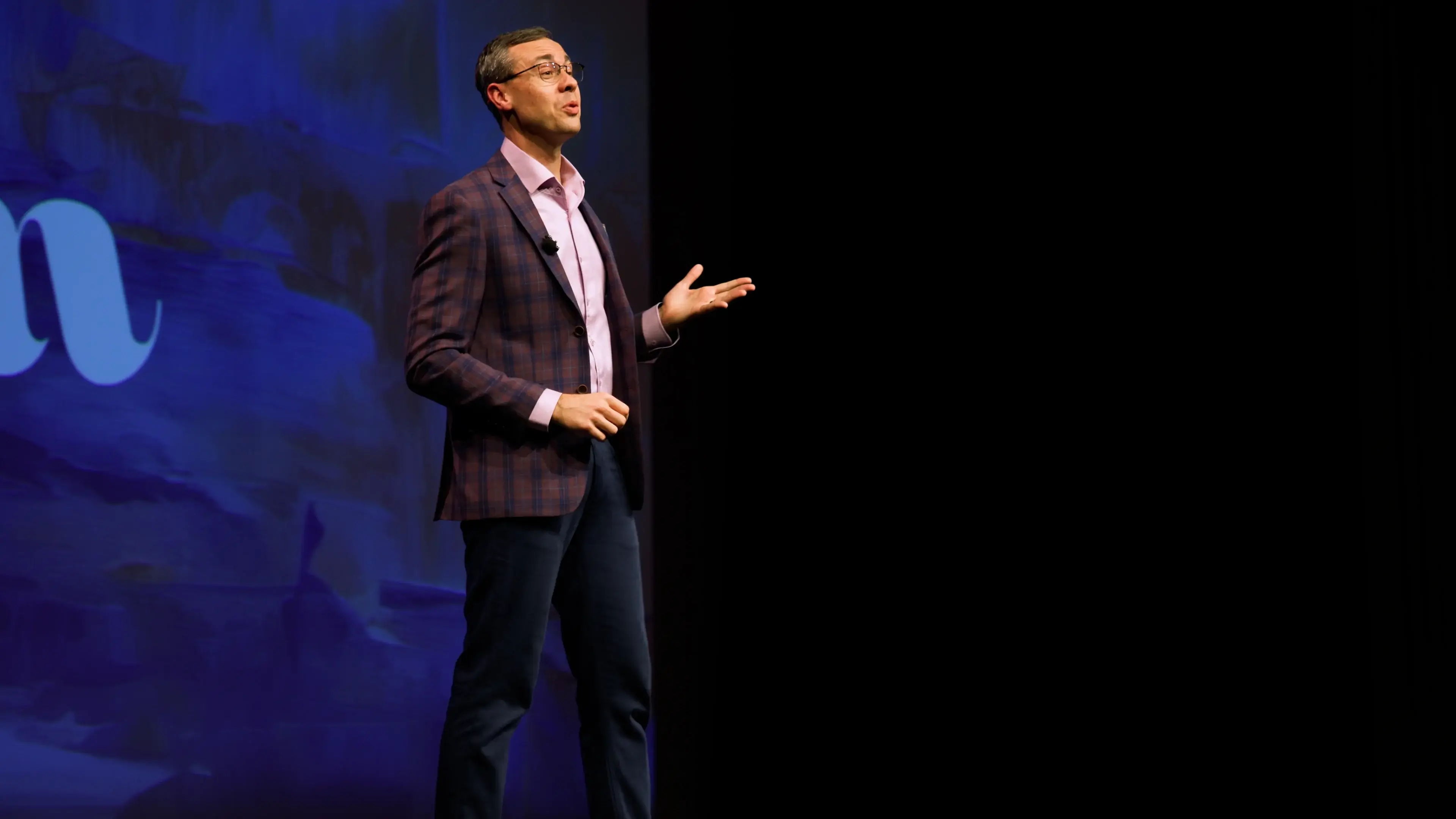 Man in glasses and a plaid blazer speaking on stage with a blue backdrop and raised hand gesture.
