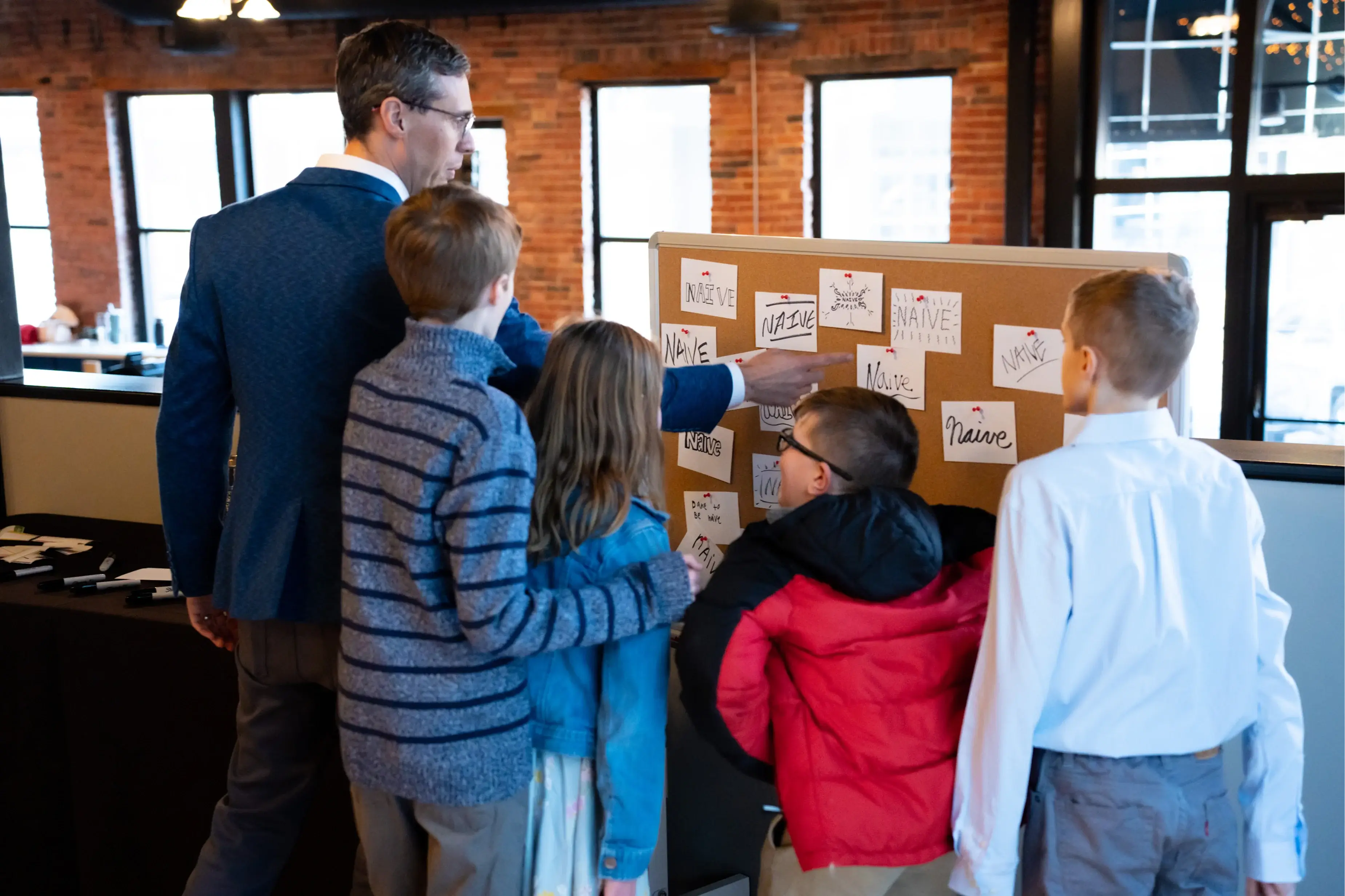 Man in a blue jacket teaching four children while pointing at a corkboard covered with notes displaying the word 'Naive' in various styles.