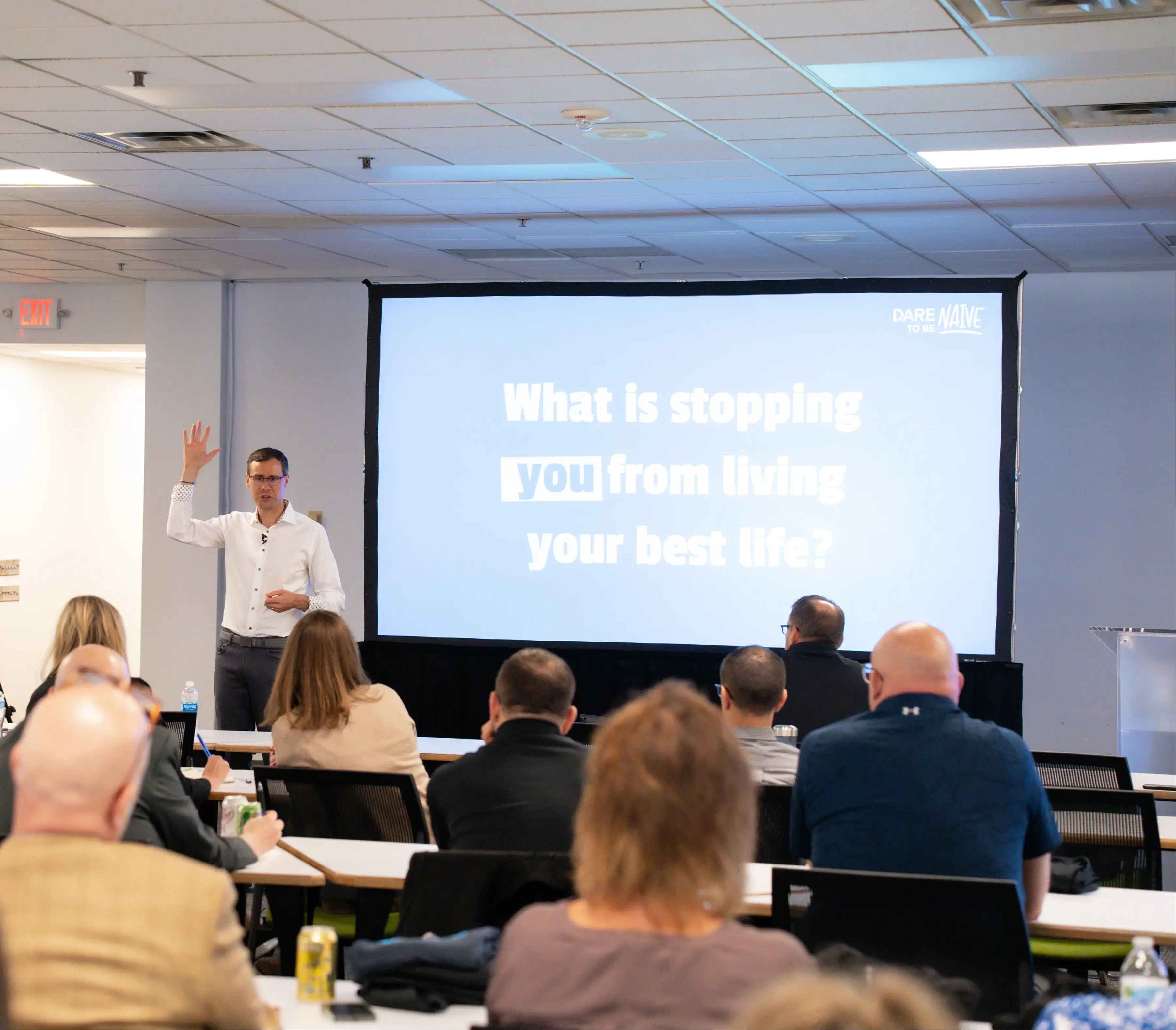 Speaker in white shirt raising hand in front of seated audience and a projector screen displaying the question, 'What is stopping you from living your best life?'