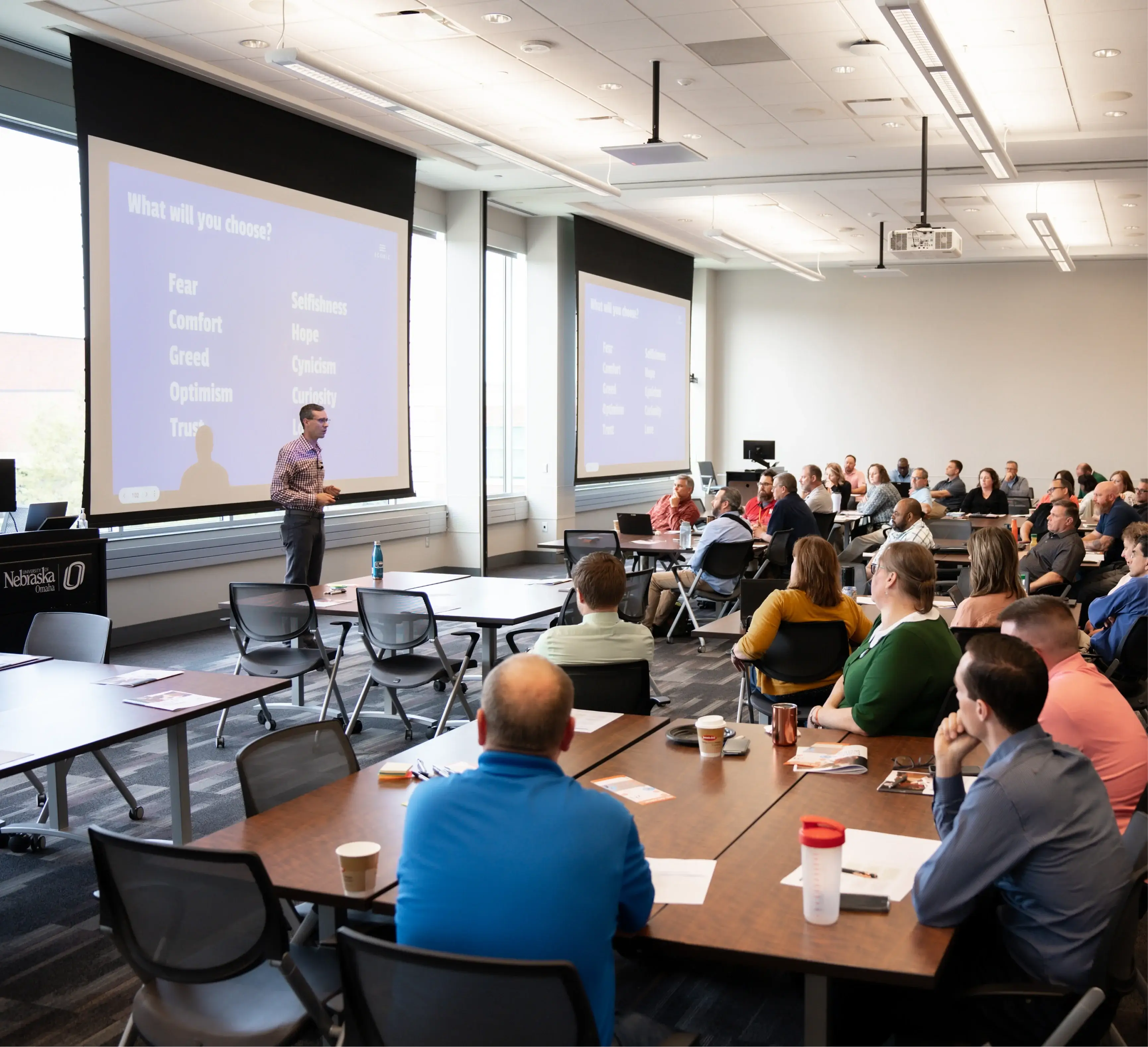 A man giving a presentation in a classroom with attendees seated at tables watching and listening.