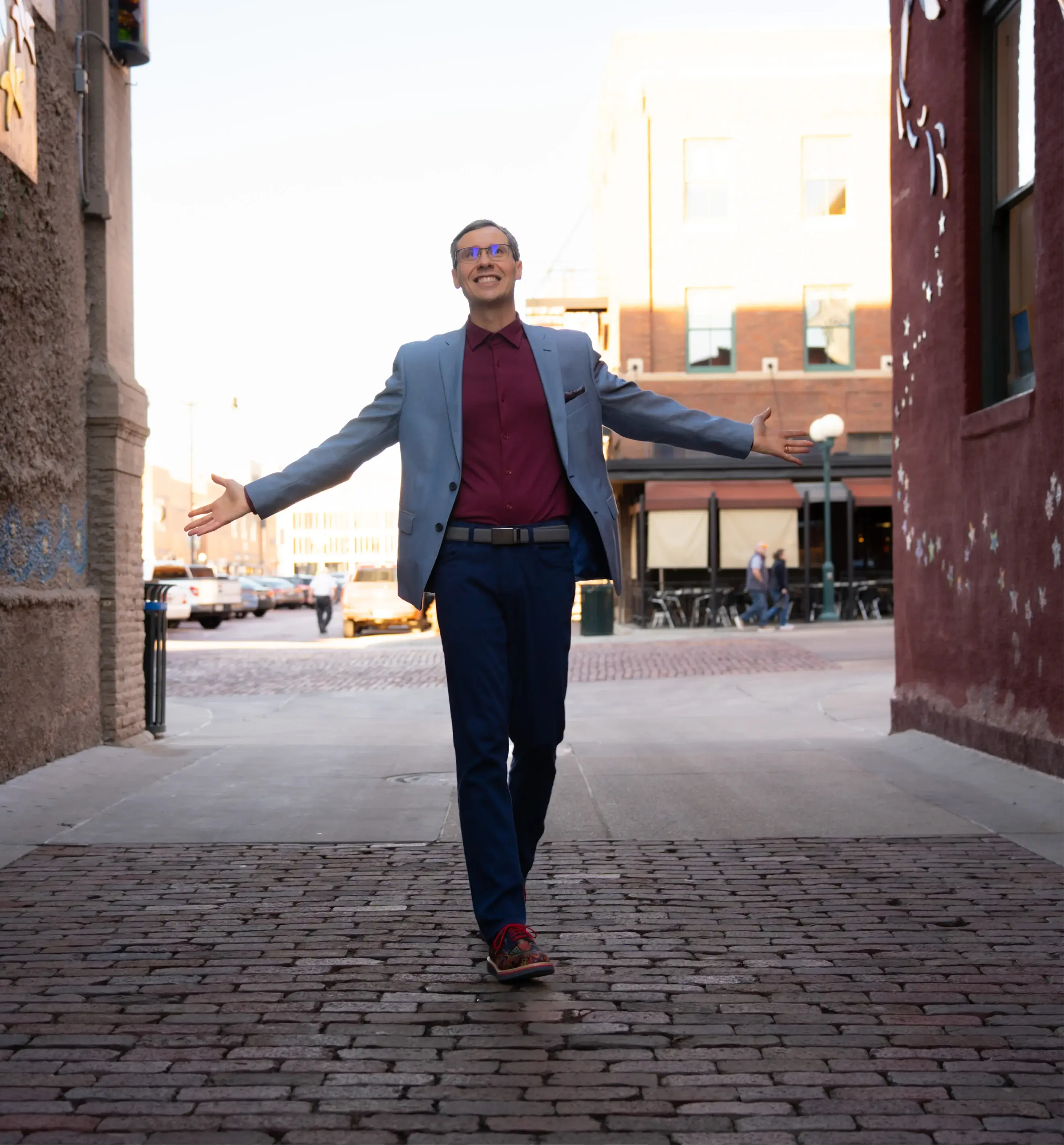 Man in a light blue blazer and burgundy shirt walking on cobblestone street with arms open wide, smiling.