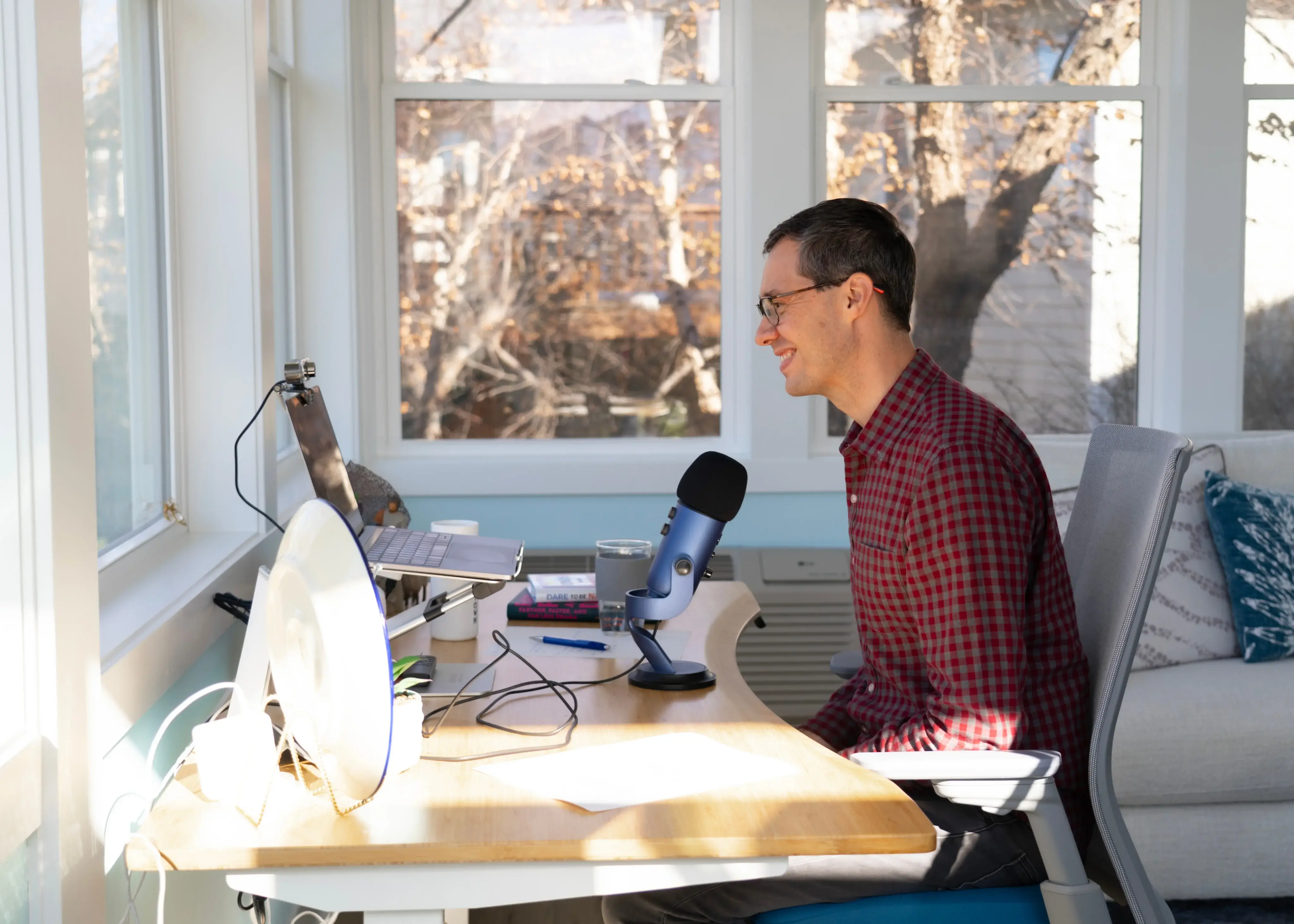 Man in glasses and red checkered shirt speaking into a microphone at a desk with a laptop and webcam in a sunlit room.