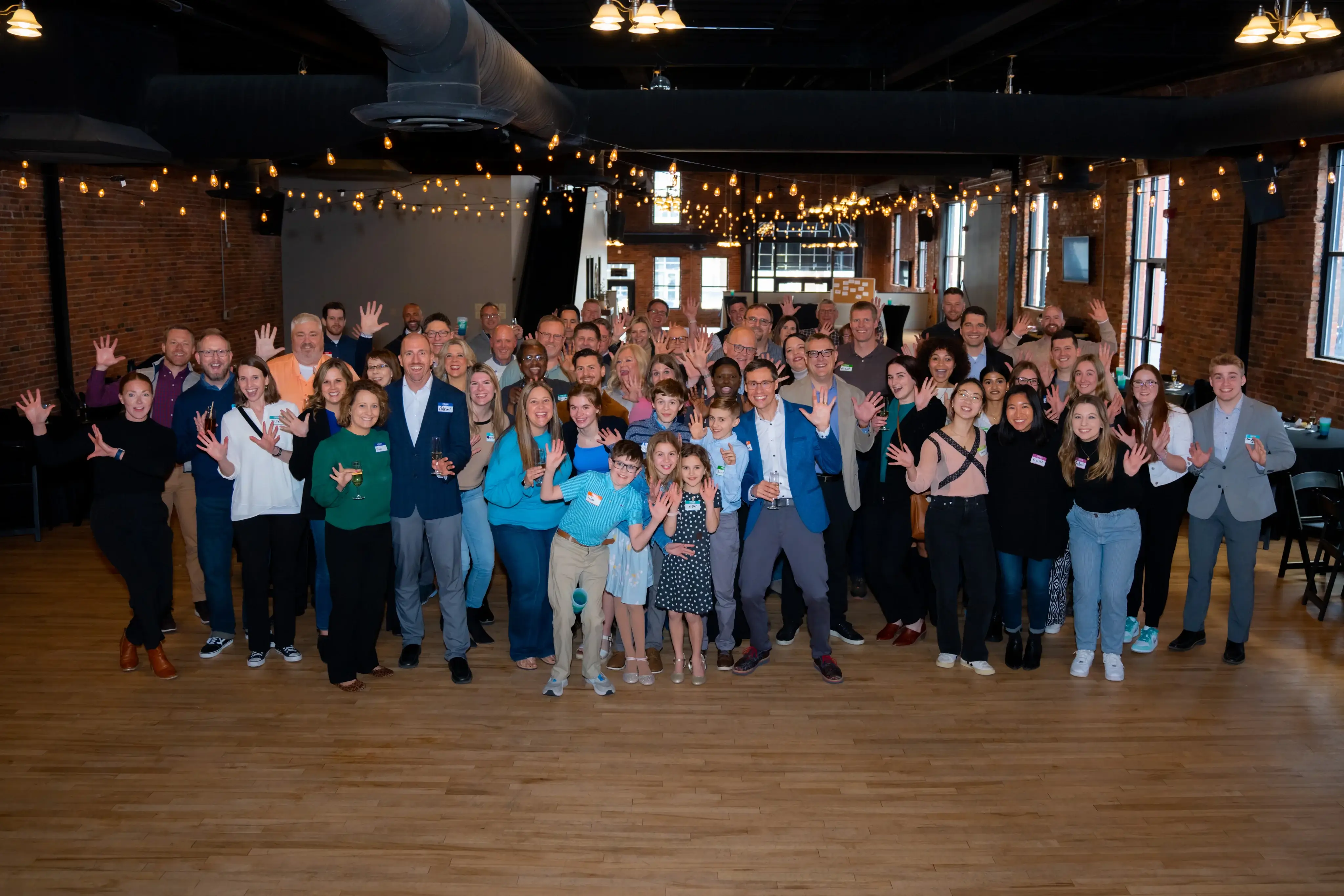 Large group of diverse people smiling and waving indoors under string lights in a spacious room with wooden floors and brick walls.