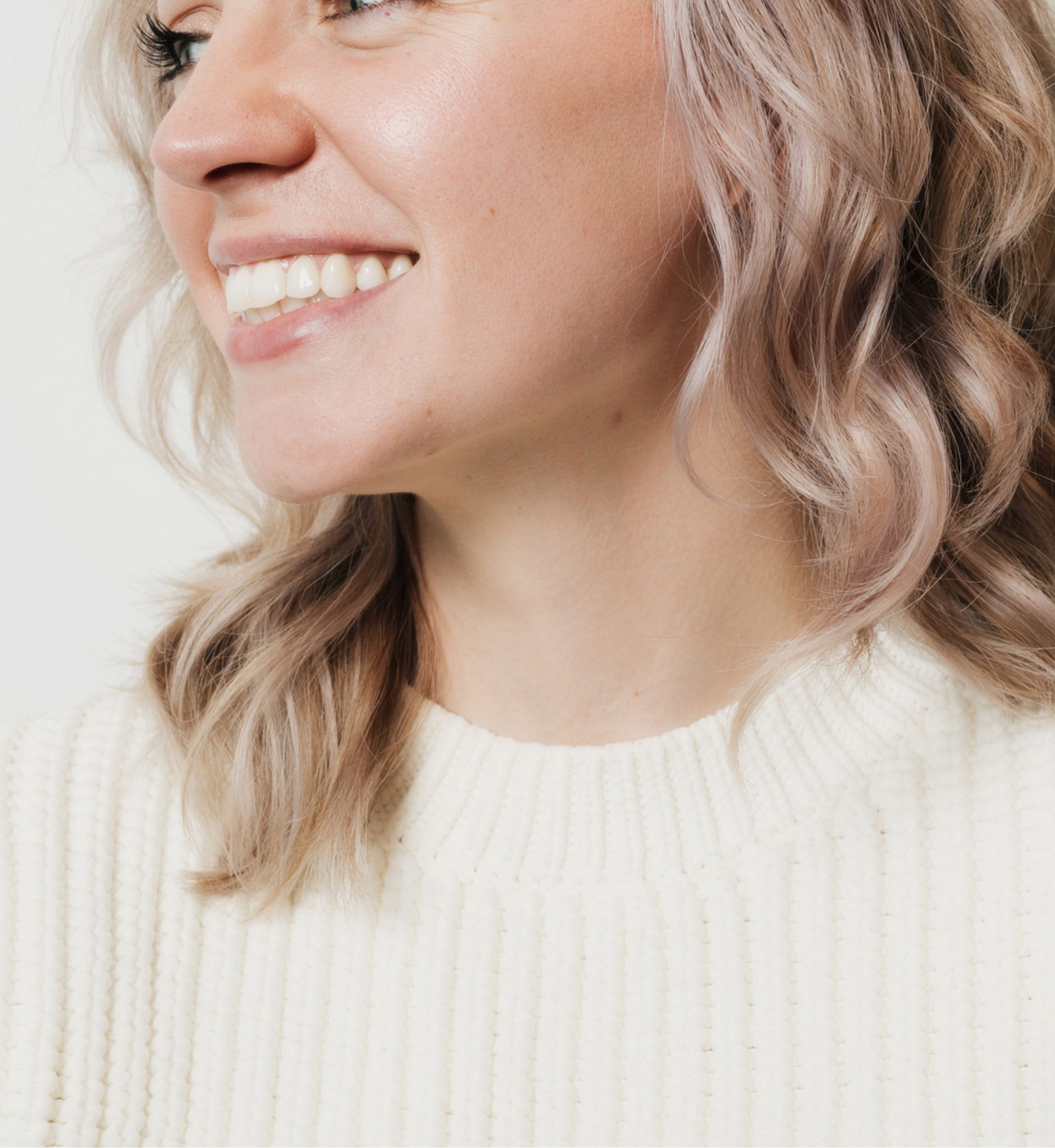 Close-up of a smiling woman with light wavy hair wearing a white knit sweater.