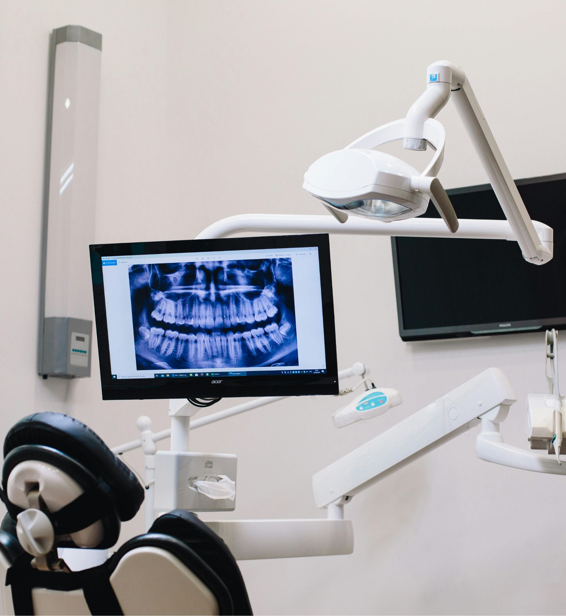 Dental examination room with a chair, an overhead dental light, and a screen displaying a panoramic dental X-ray.