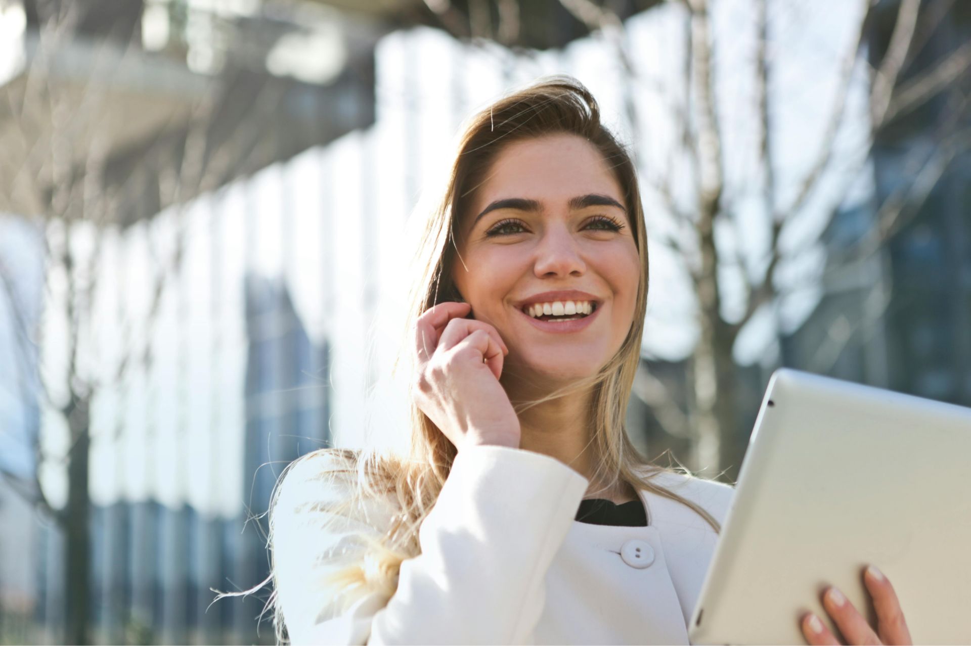 Smiling woman outdoors holding a tablet and touching her hair.