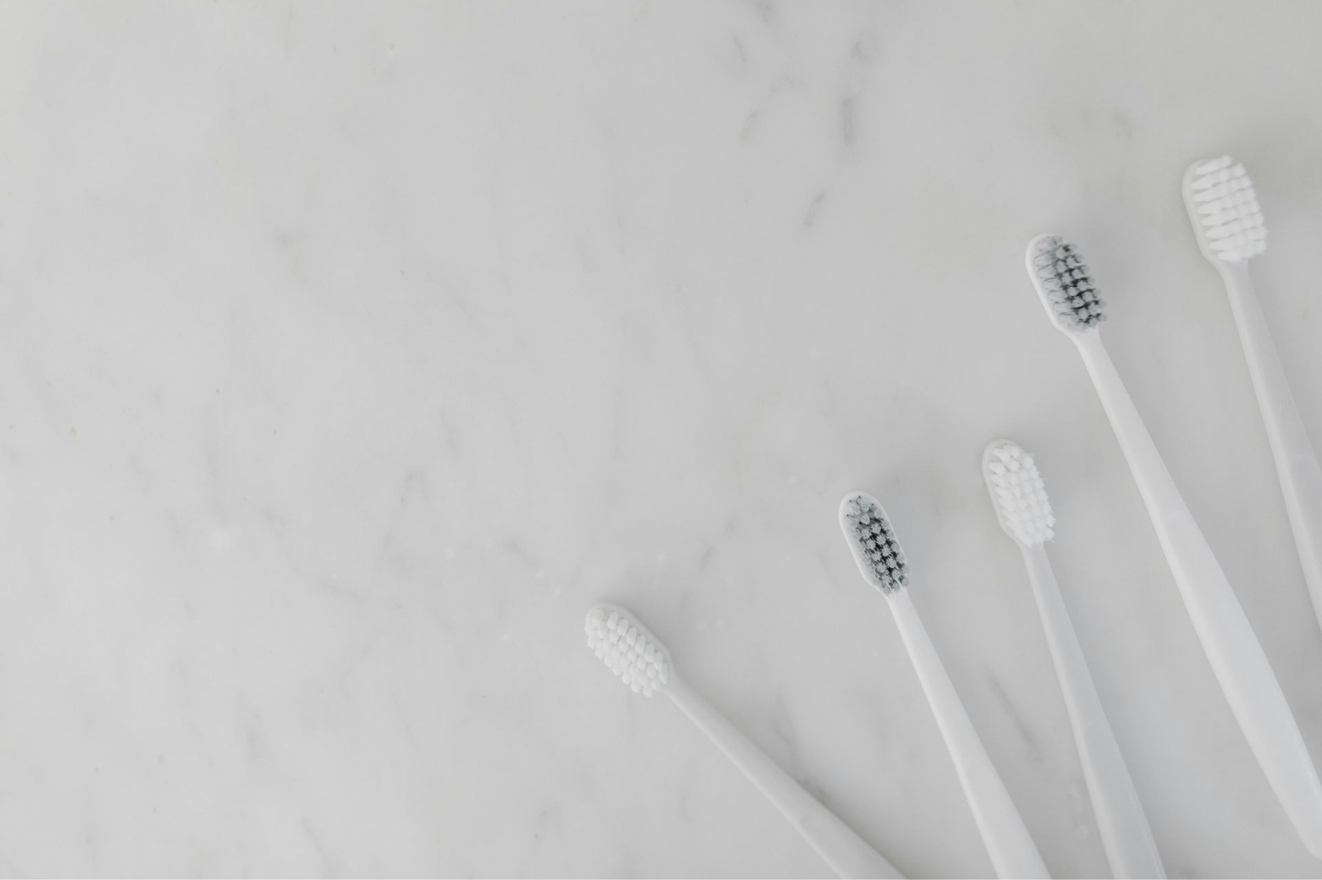 Five white toothbrushes with some having black bristles arranged fanned out on a light marble surface.