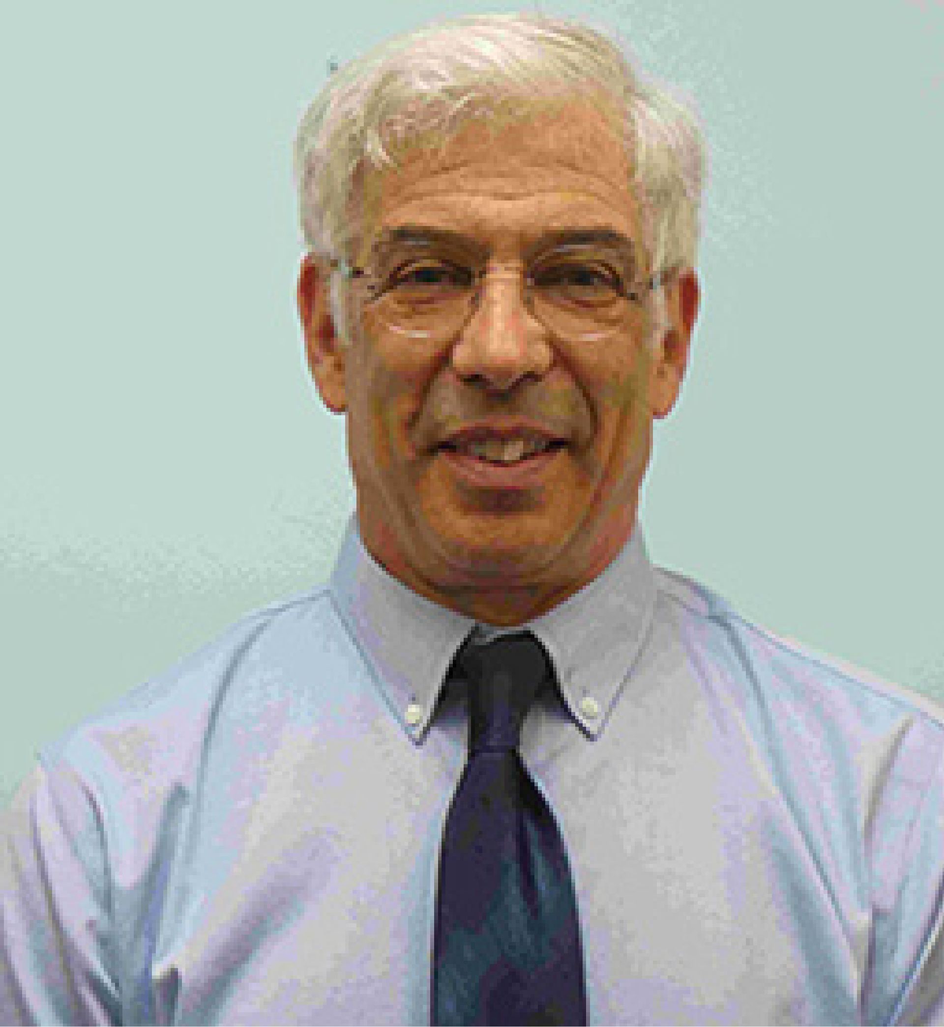 Smiling elderly man with white hair and glasses wearing a light blue shirt and dark tie against a plain light background.