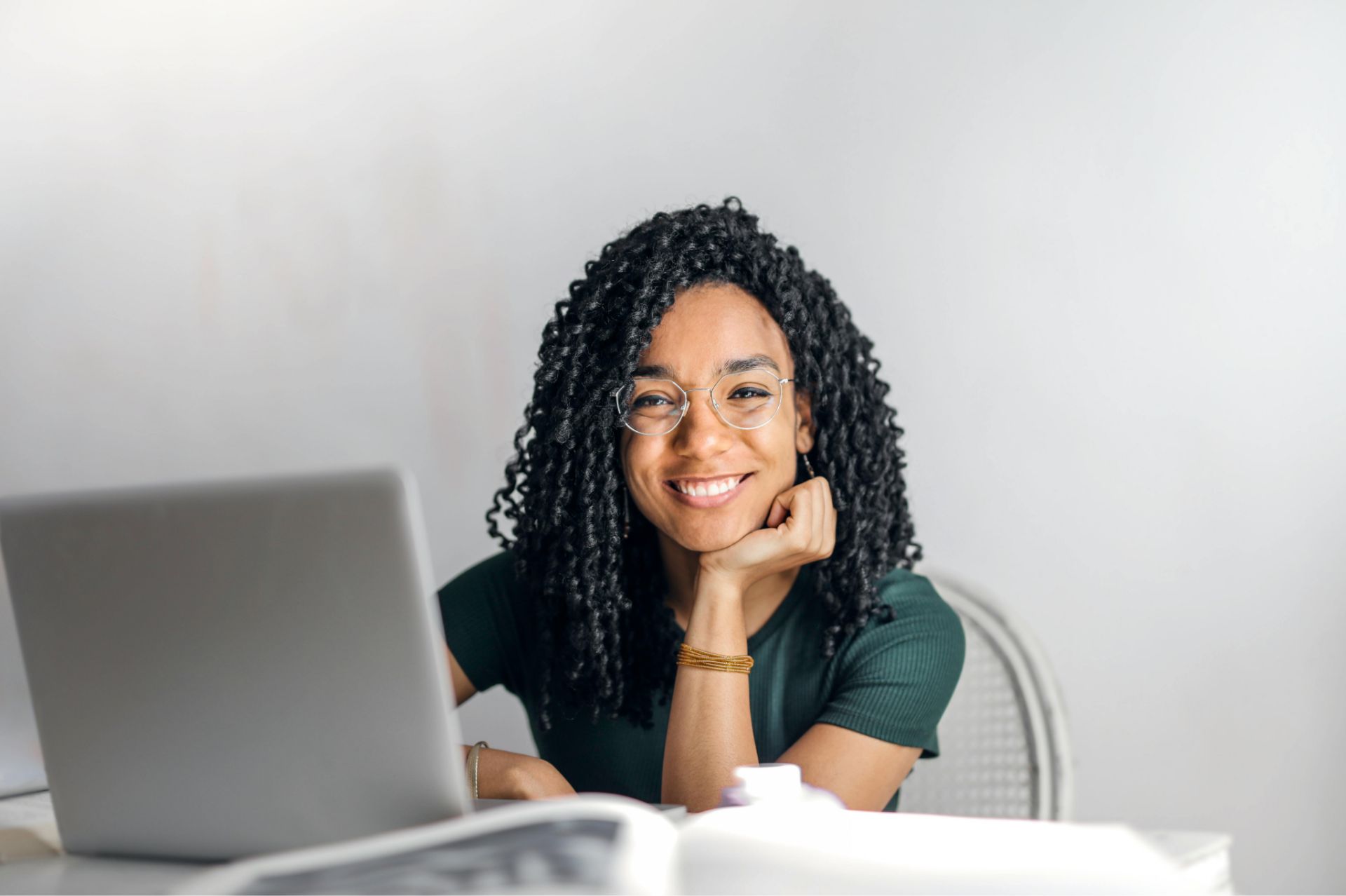 Smiling woman with curly hair and glasses working on a laptop at a bright desk.