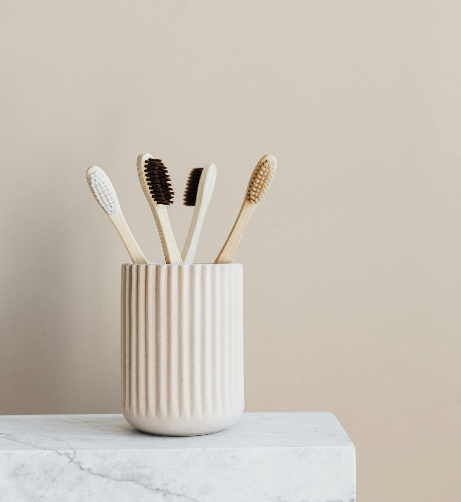 Four bamboo toothbrushes with varying bristle colors in a beige ribbed holder on a light marble surface.