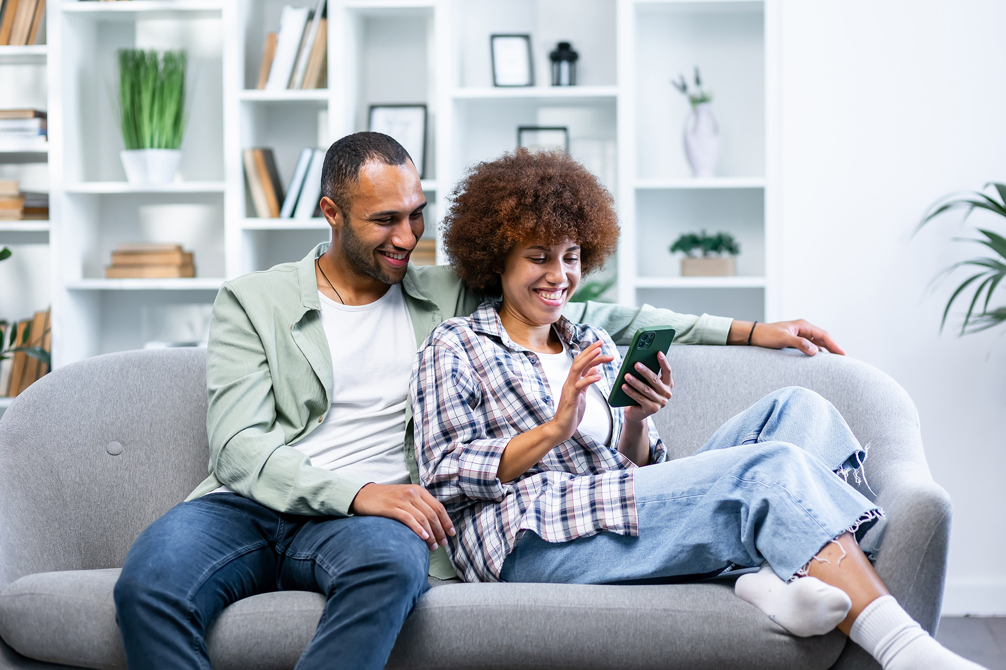 Smiling couple sitting on a gray couch looking at a smartphone together in a bright room with shelves and plants.