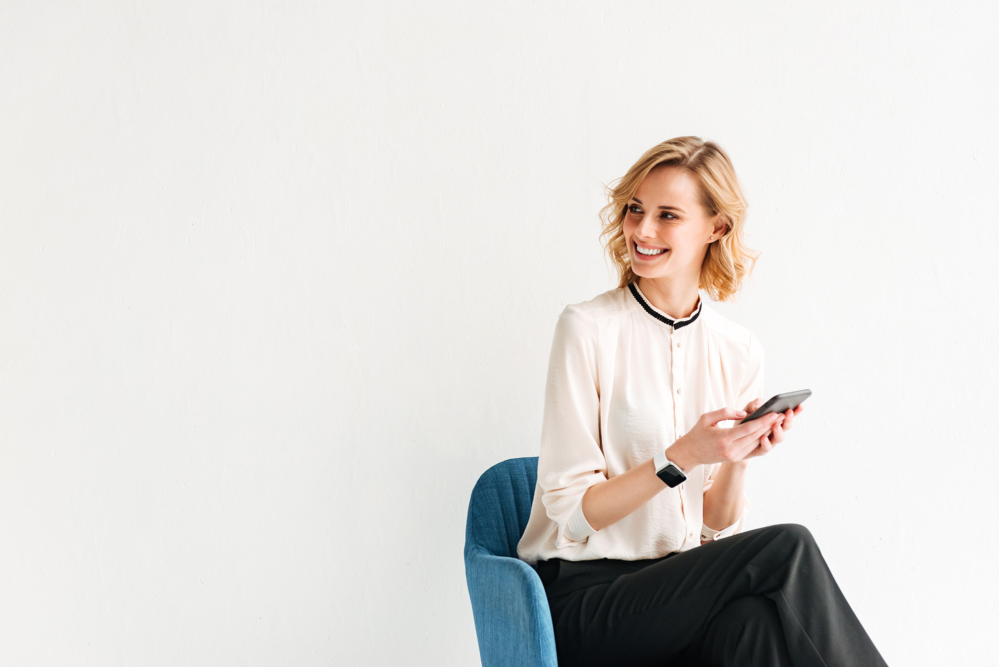 Smiling woman sitting on a blue chair using a smartphone against a white background.