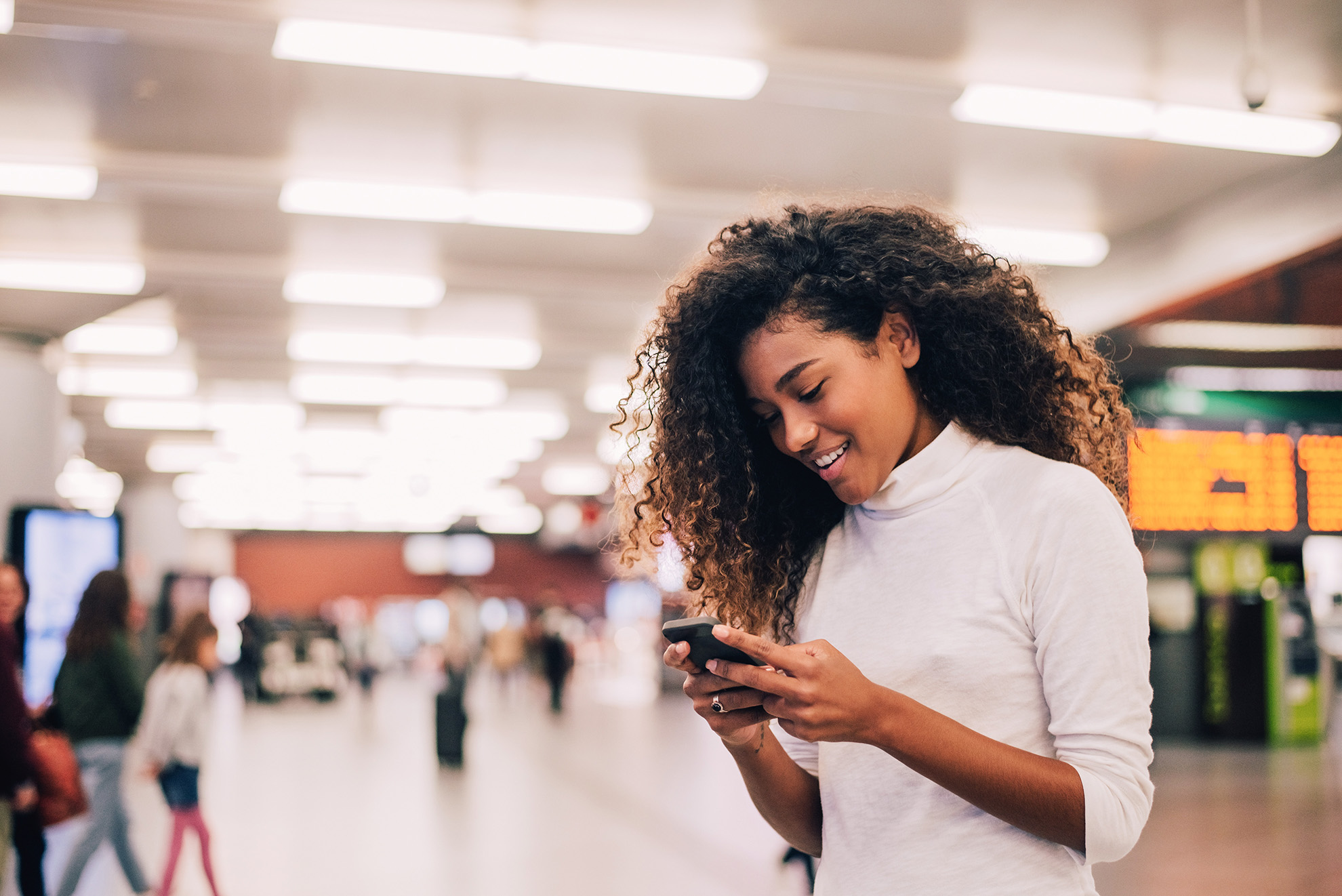 Smiling young woman with curly hair looking at her smartphone in a busy indoor public space.