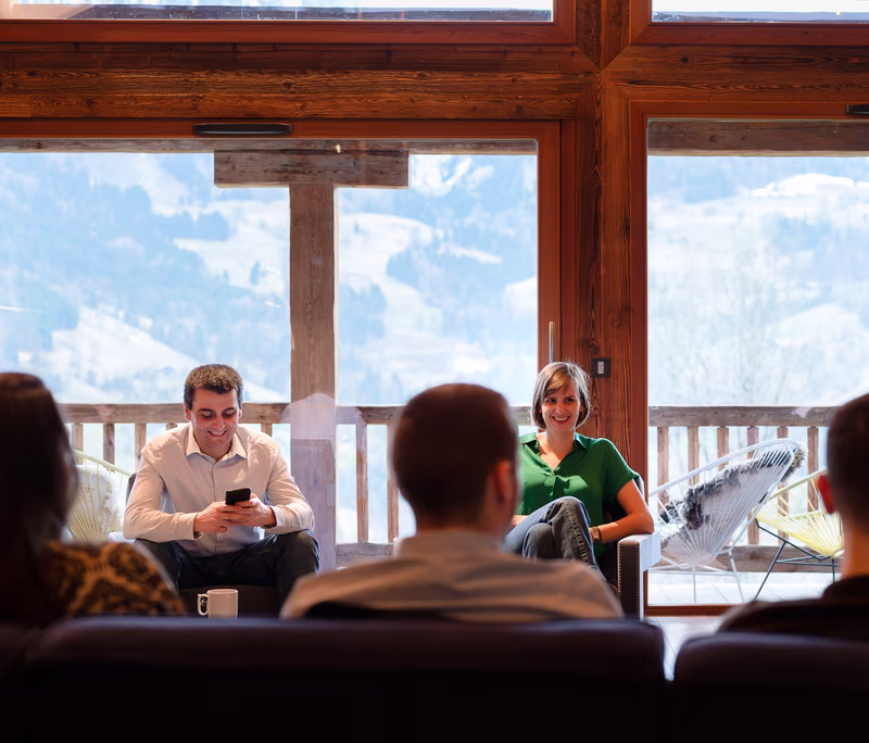 Deux personnes assises sur des fauteuils face à trois autres autour d'une table, avec une grande fenêtre montrant un paysage de montagne en arrière-plan.