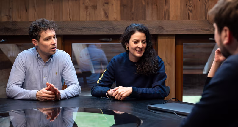 Two people sitting at a black table talking to a partially visible third person.