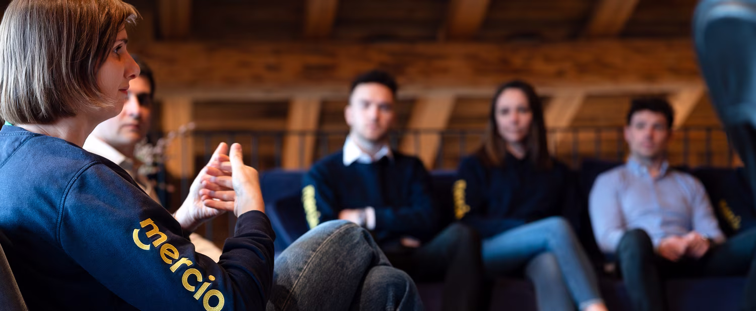 Group of five people sitting and talking in a room with wooden beams visible on the ceiling.