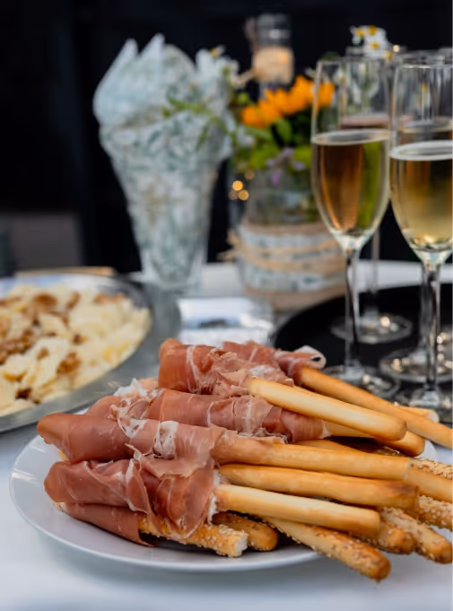 Breit angerichtete Sardellenstangen mit dünn geschnittenem Prosciutto auf weißem Teller, im Hintergrund Champagnergläser und Blumen.