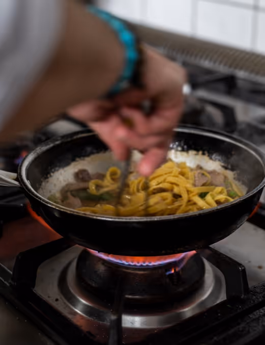 Person rührt Nudeln und Pilze in einer Pfanne auf einem Gasherd.