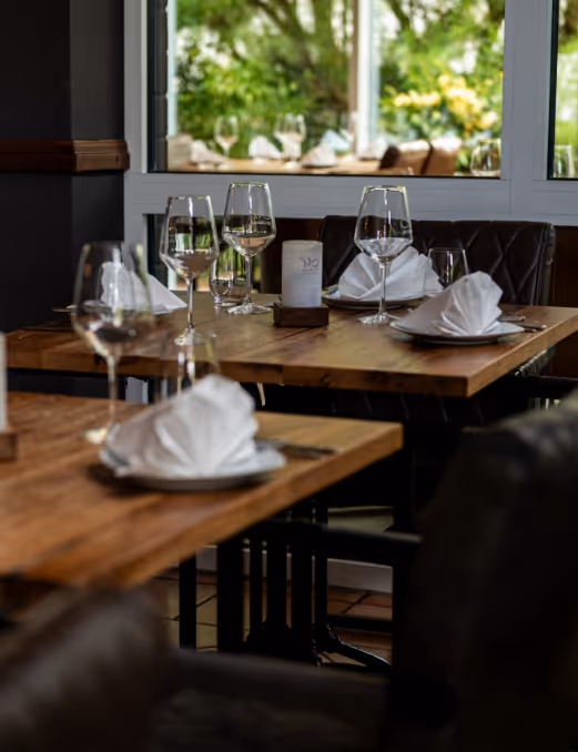 Eleganter gedeckter Tisch in einem Restaurant mit gefalteten Servietten und Weingläsern vor einem Fenster mit Blick auf grüne Pflanzen.