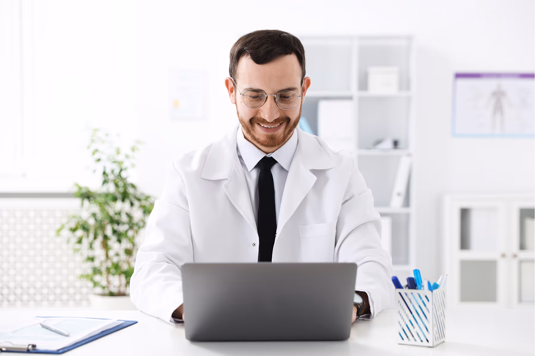Smiling male doctor with glasses working on a laptop at a white desk in a bright office.