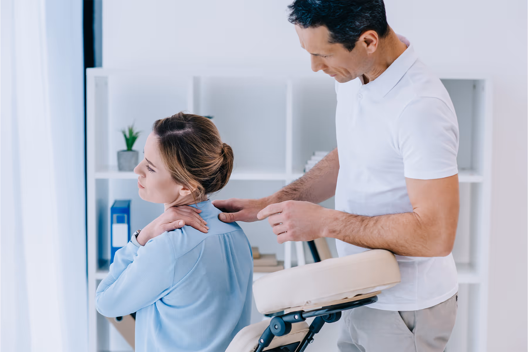 Woman in blue shirt holding her neck in pain while a man in white shirt examines her shoulder in a bright room.