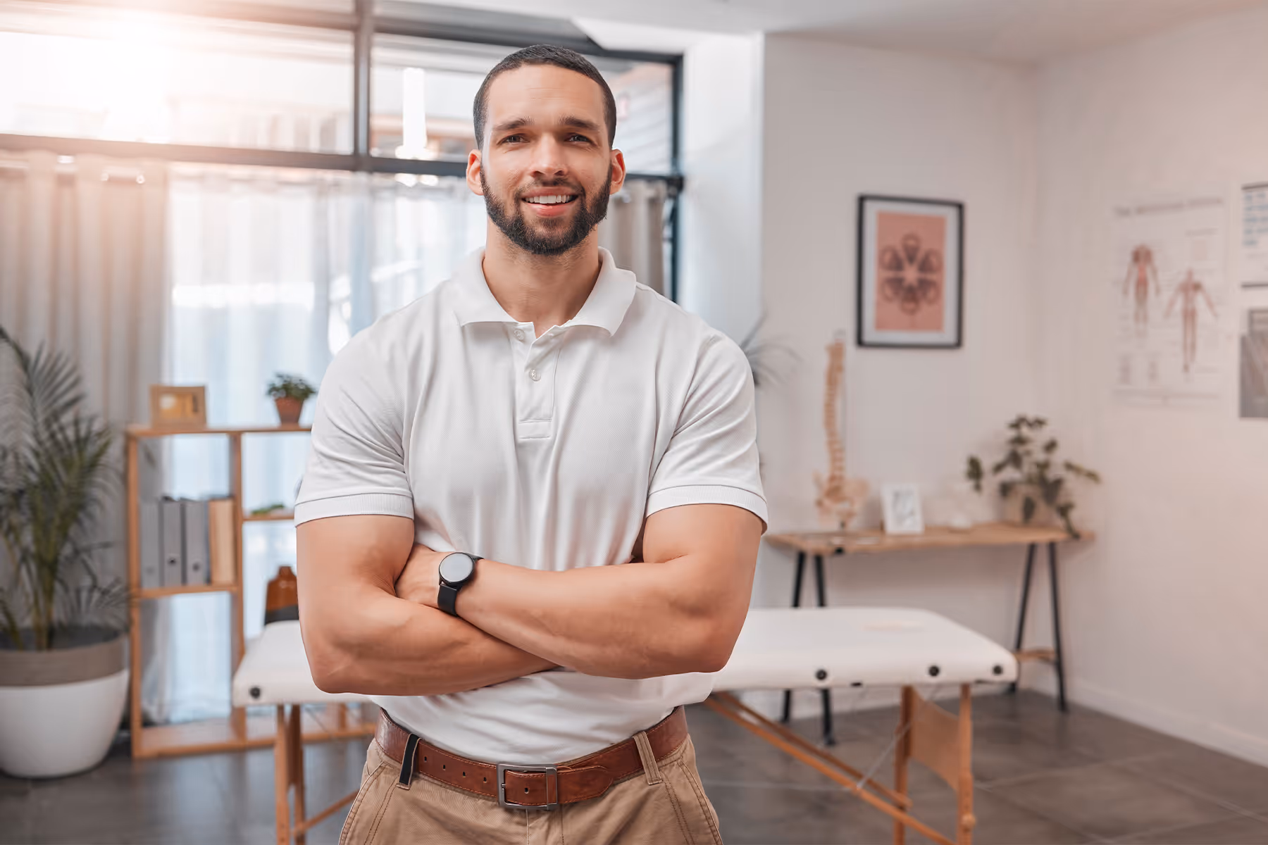 Smiling man in a white polo shirt stands with arms crossed in a bright medical or therapy room.