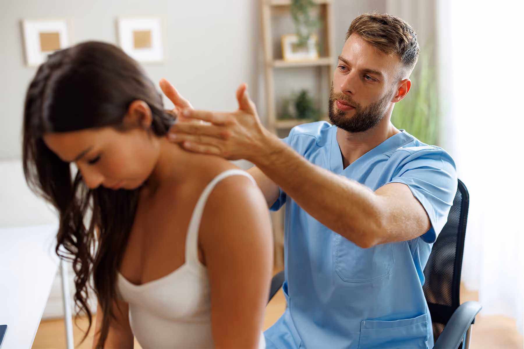Male physical therapist in blue scrubs examining a young woman's neck in a bright room.