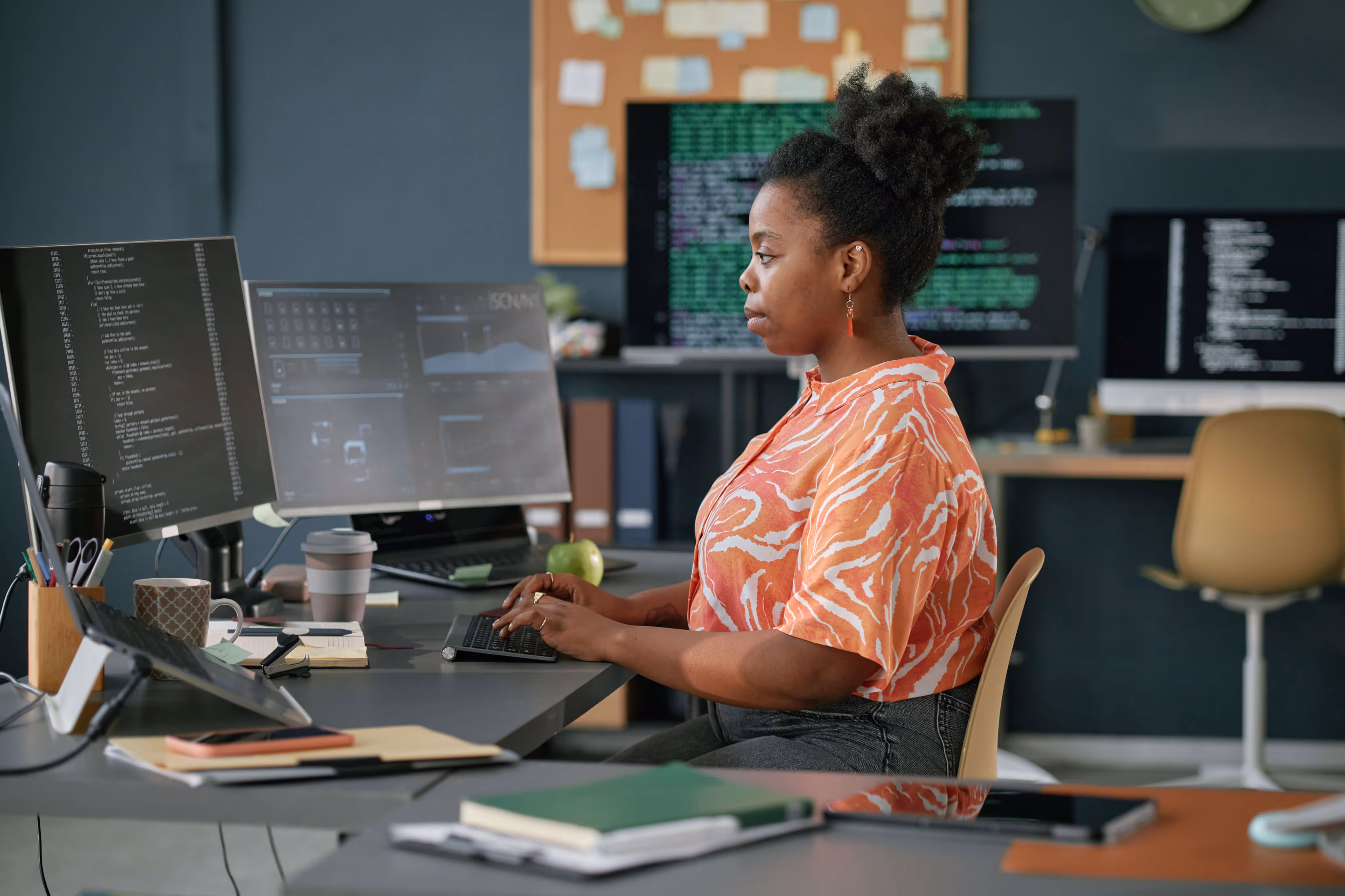 Woman in an orange patterned shirt typing on a keyboard at a desk with multiple computer monitors displaying code and data.