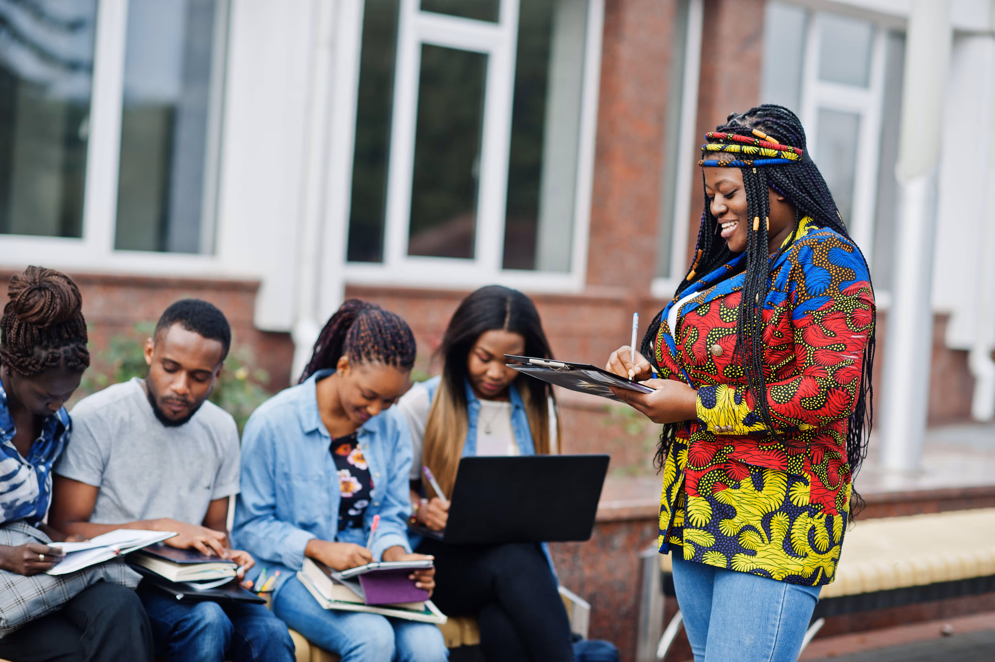 A group of young adults studying outdoors with notebooks and a laptop, one woman in colorful patterned clothing standing and writing on a clipboard.