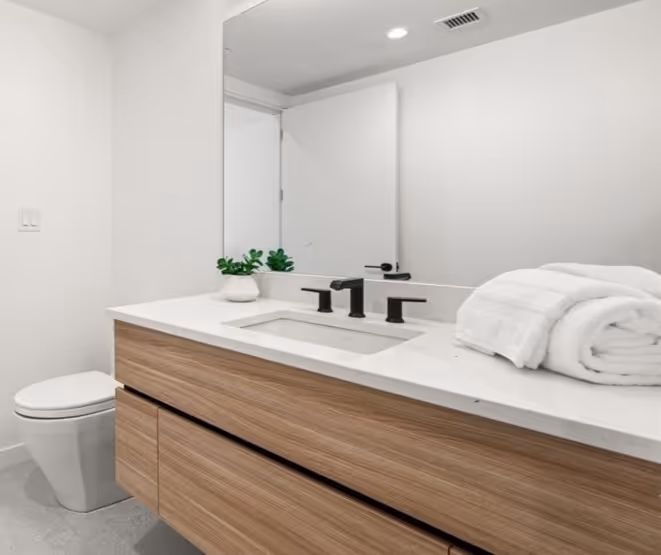 Modern bathroom with wooden vanity, white countertop, black faucet, small plant, rolled white towel, and white toilet.
