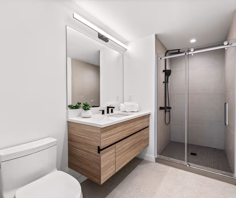 Modern bathroom with floating wooden vanity, large mirror with light, white toilet, and a glass sliding door shower with black fixtures.