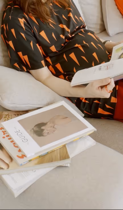 Pregnant woman in a black dress with orange patterns sitting on a couch reading a book, with several books on the table nearby.