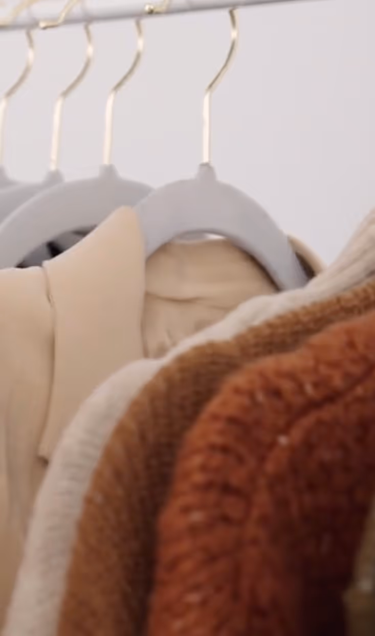 Close-up of neutral-toned sweaters and coats hanging on a clothing rack with gray hangers.