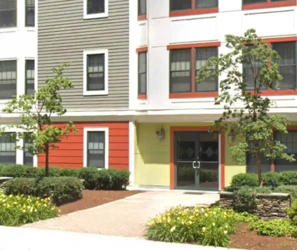 Modern apartment building entrance with a walkway, small trees, and landscaped bushes and flowers.