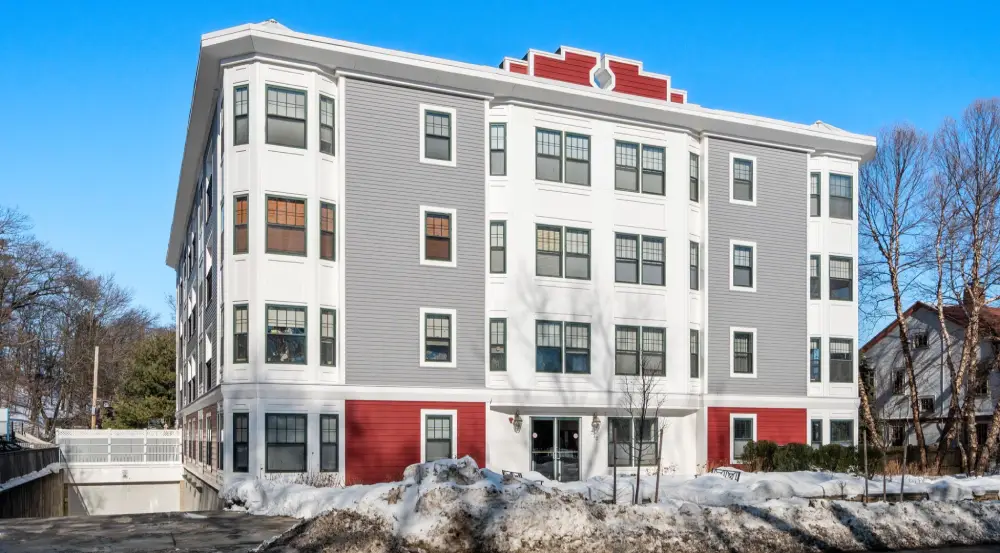 Four-story apartment building with white and gray siding, red accents, and snow-covered ground in front.