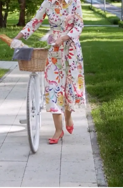 Person wearing a colorful floral dress and red shoes walking a white bicycle with a wicker basket on a paved path beside green grass.