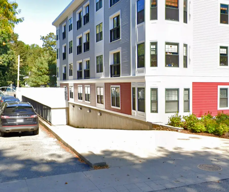 Multi-story residential building with a sloped driveway leading to an underground parking entrance and several parked cars alongside.