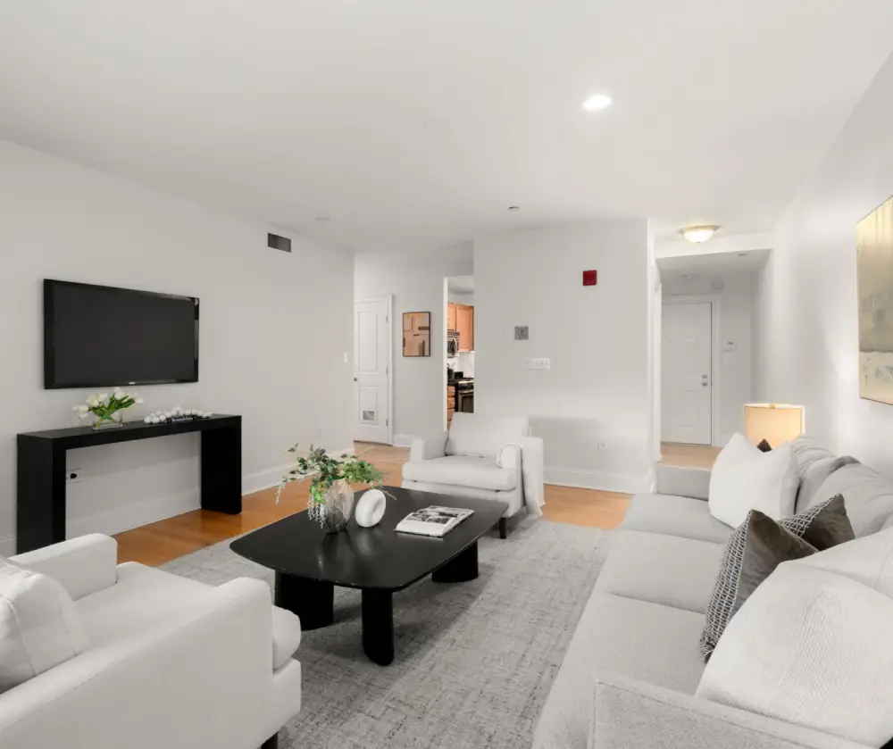 Modern living room with white walls, gray sofa, two white armchairs, black coffee and TV tables, and wooden floor.