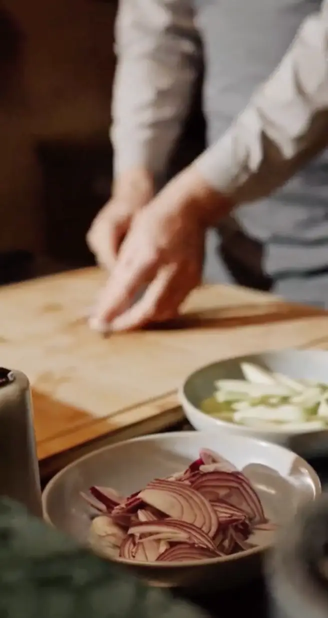 Person chopping ingredients on a wooden cutting board with sliced onions and other vegetables in bowls nearby.
