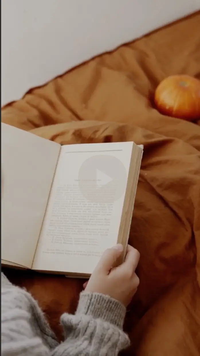 Person wearing a gray sweater reading an open book on a brown blanket with a small pumpkin nearby.
