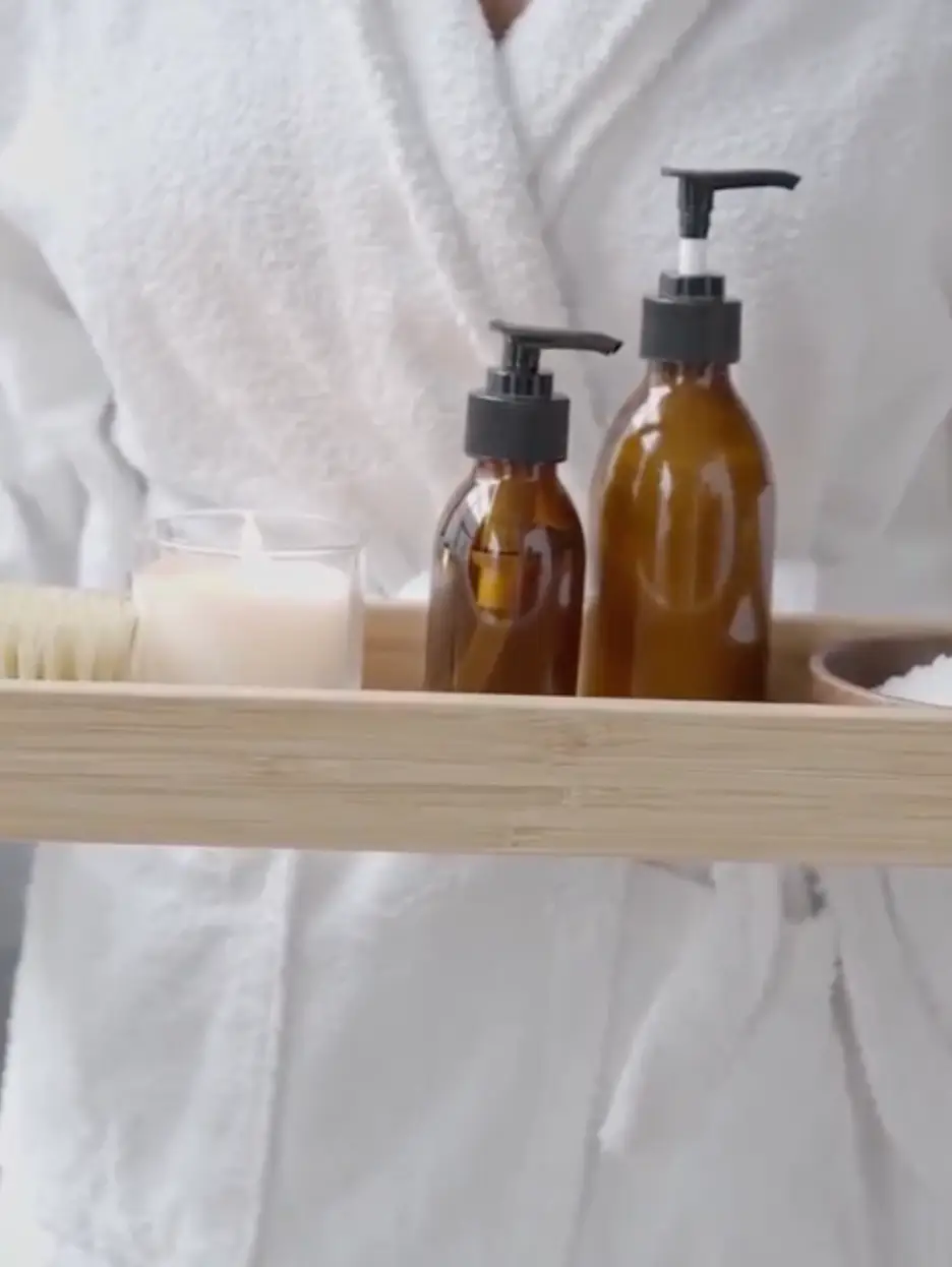 Person in white bathrobe holding a wooden tray with amber pump bottles, a candle, a brush, and a bowl with bath salts.
