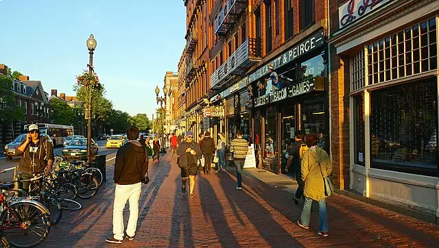 People walking on a sunlit brick sidewalk lined with bicycles and red brick buildings in a city street scene.
