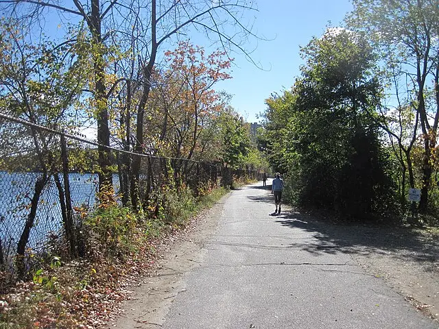 Person walking on a paved path alongside a chain-link fence with trees showing autumn foliage and a body of water visible through the fence under a clear blue sky.