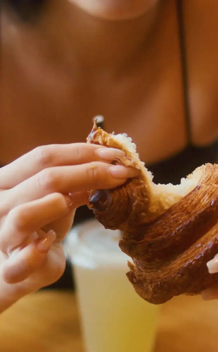 Person with manicured nails tearing apart a chocolate croissant with a blurred drink in the background.
