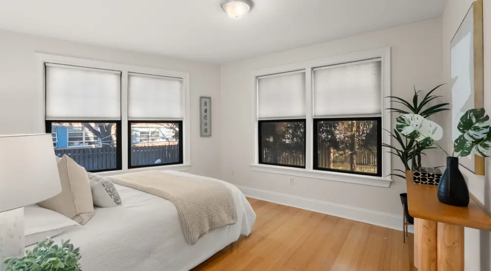 Bright bedroom with two sets of windows with white blinds, a bed with beige and white bedding, wooden floor, and a wooden table with plants and a framed picture.