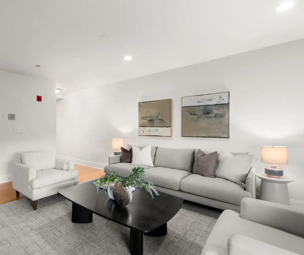 Modern living room with gray sofa, two armchairs, black coffee table with vase and greenery, two abstract paintings, and side tables with lamps.