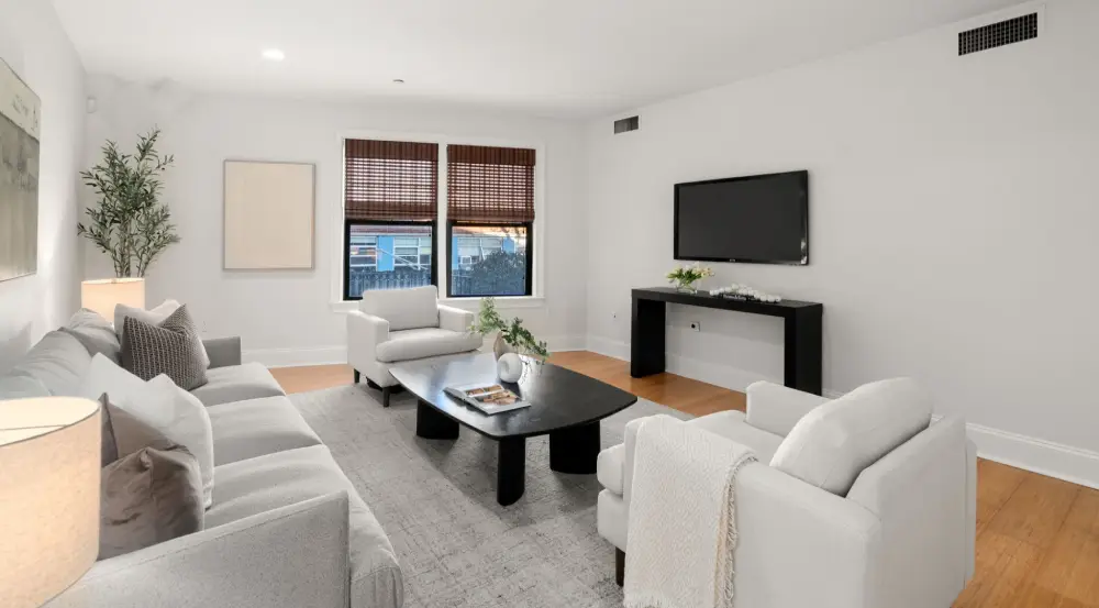 Modern living room with white sofas, black coffee table, wall-mounted flat screen TV, and two windows with wooden blinds.