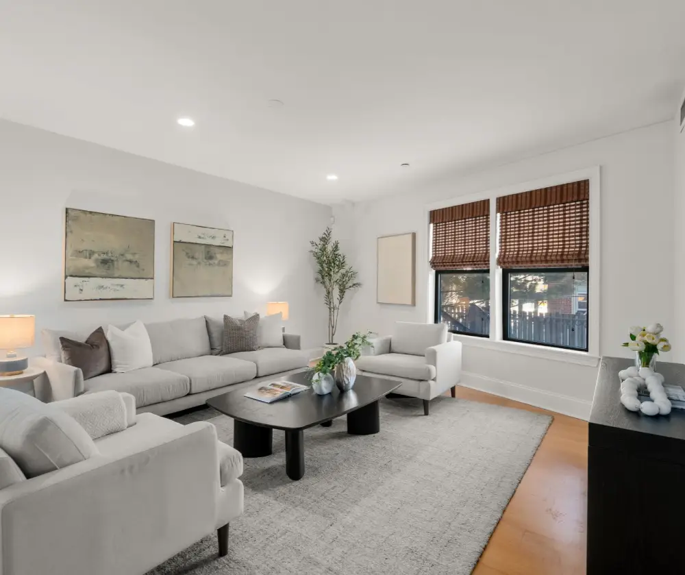 Modern living room with a light gray sofa, two armchairs, a black coffee table with decorative vases, and two windows with brown blinds.